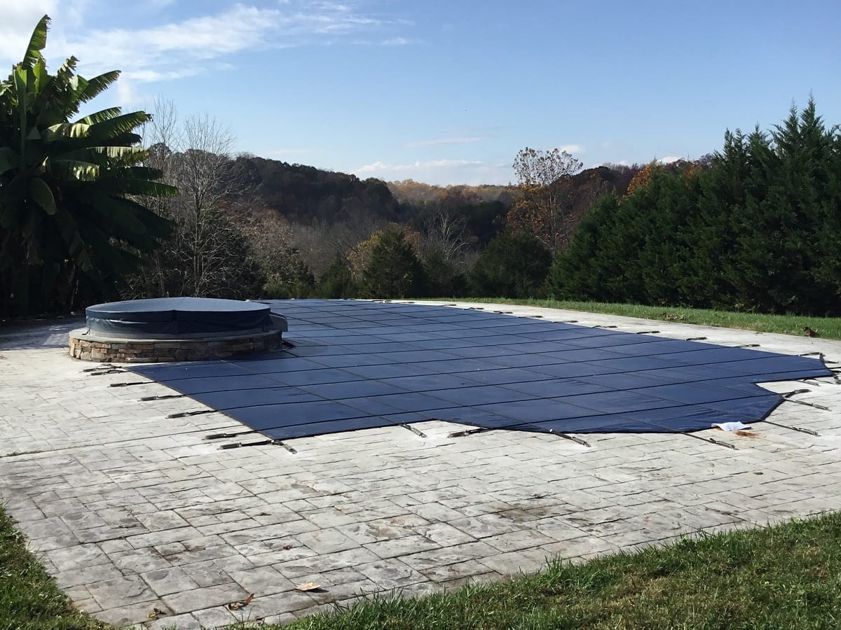 Blue mesh safety cover installed on an inground pool with a raised spa, fall hills behind